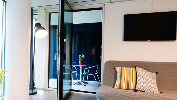 A modern living room with a sofa, a flat-screen TV, and a glass door leading to a balcony with a table and chairs. - Apartment  by Interhome (Lucerne)