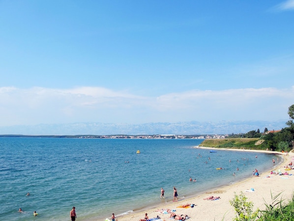 Ein Strand mit badenden und sonnenbadenden Personen, ein klarer Himmel und Berge im Hintergrund.