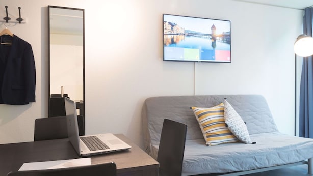 Ein modernes Hotelzimmer mit Schreibtisch, Laptop und einer grauen Couch mit Zierkissen. Ein Flachbildfernseher ist an der Wand befestigt.