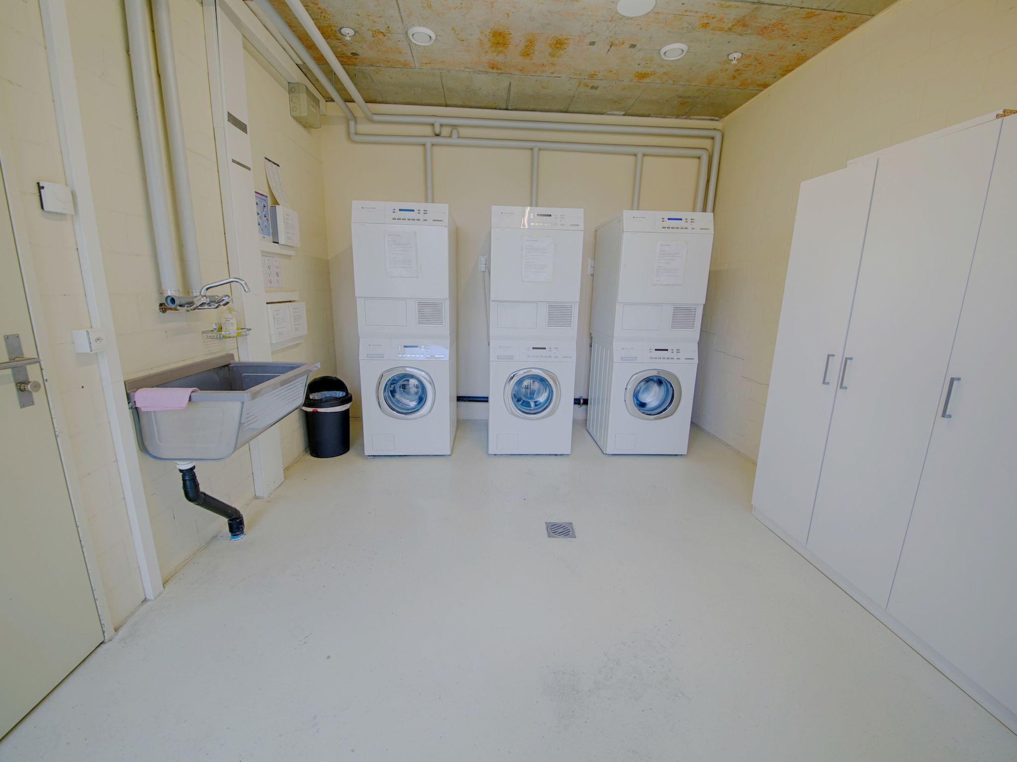 A laundry room with three stacked washing machines, a sink, and a trash can.