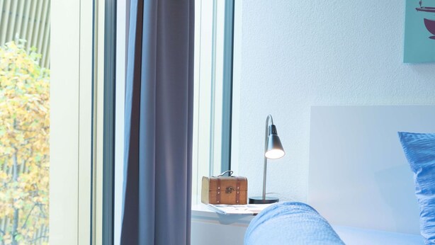 A bed with a headboard, a bedside lamp, a small wooden box, and a window with curtains. - Apartment  by Interhome (Lucerne)