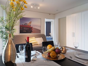 Habitación de hotel con mesa de comedor para dos, una botella de vino, frutero y cama con vista a la playa.