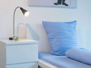 Un lit bien fait, avec un oreiller bleu et une table de chevet blanche ornée d’une lampe et d’un vase en verre.