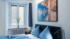 Habitación de hotel con cama, mesita de noche, lámpara, ventana con persianas, radiador y un cuadro en la pared.
