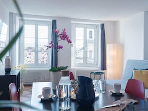 Habitación de hotel moderna con una mesa de comedor, sillas, un florero con flores, una botella de agua y vistas a la ciudad a través de amplios ventanales.