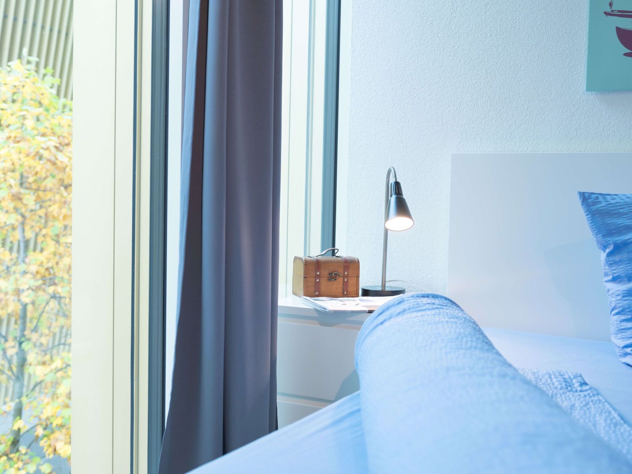 A bed with a headboard, a bedside lamp, a small wooden box, and a window with curtains.