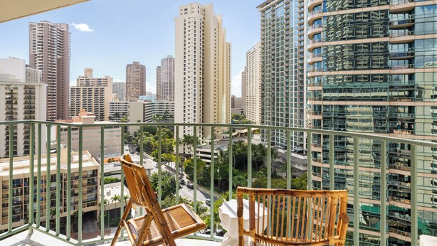 A balcony with two wicker chairs, overlooking a cityscape with high-rise buildings and a clear sky. - Delightful 2-bedroom apartment with AC in vibrant Honolulu (Honolulu)