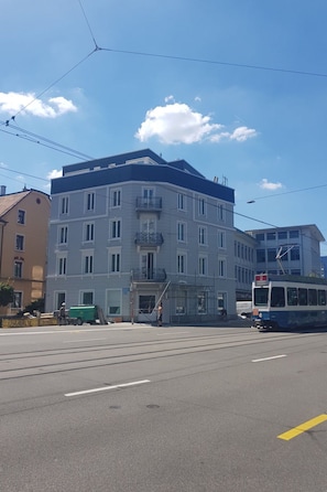 Ein mehrstöckiges Gebäude mit einem Balkon im zweiten Stock, eine Straßenbahn im Vordergrund und ein klarer Himmel.