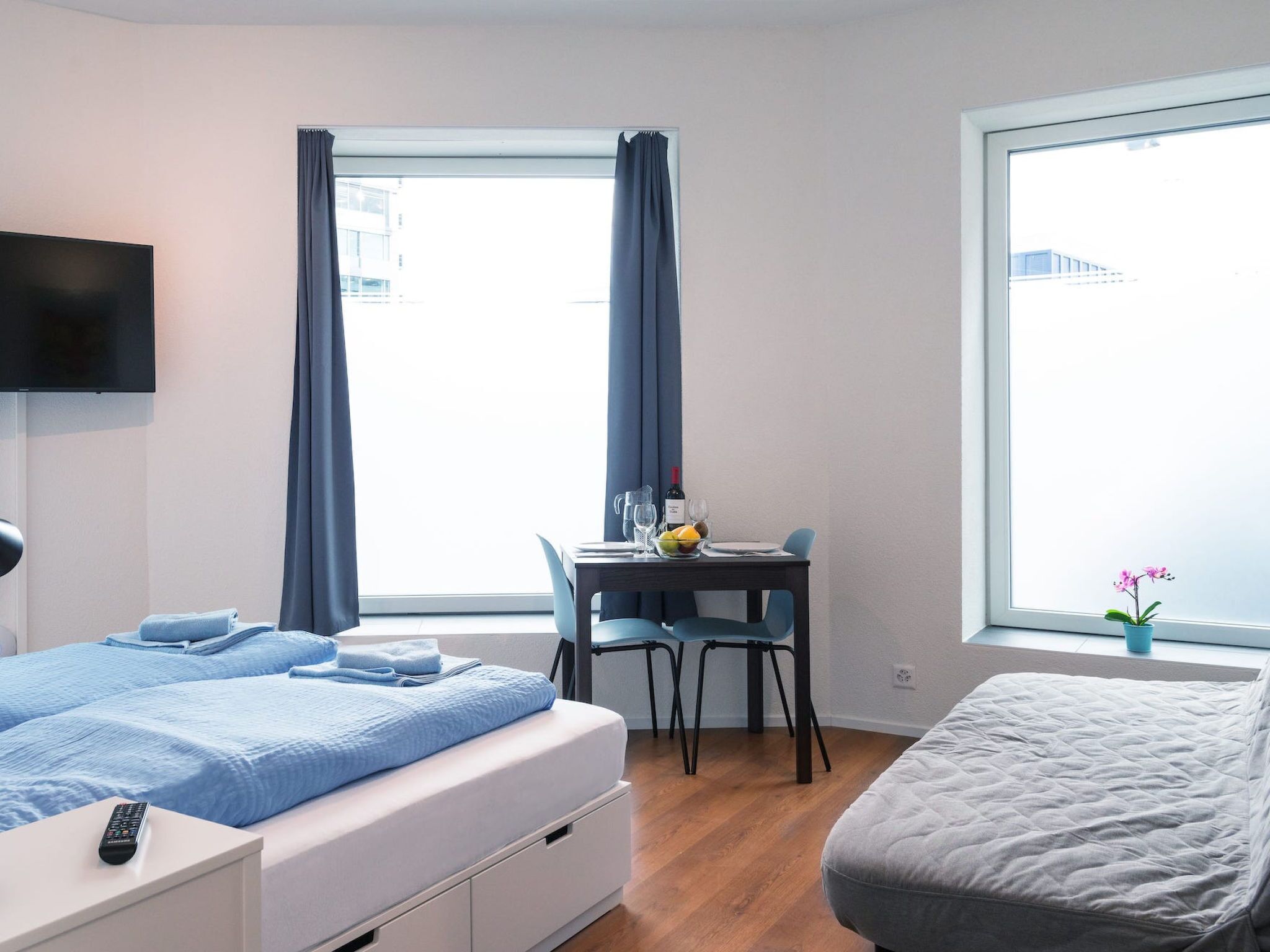 A modern hotel room with a bed, a desk with a chair, a TV mounted on the wall, and a window with curtains.