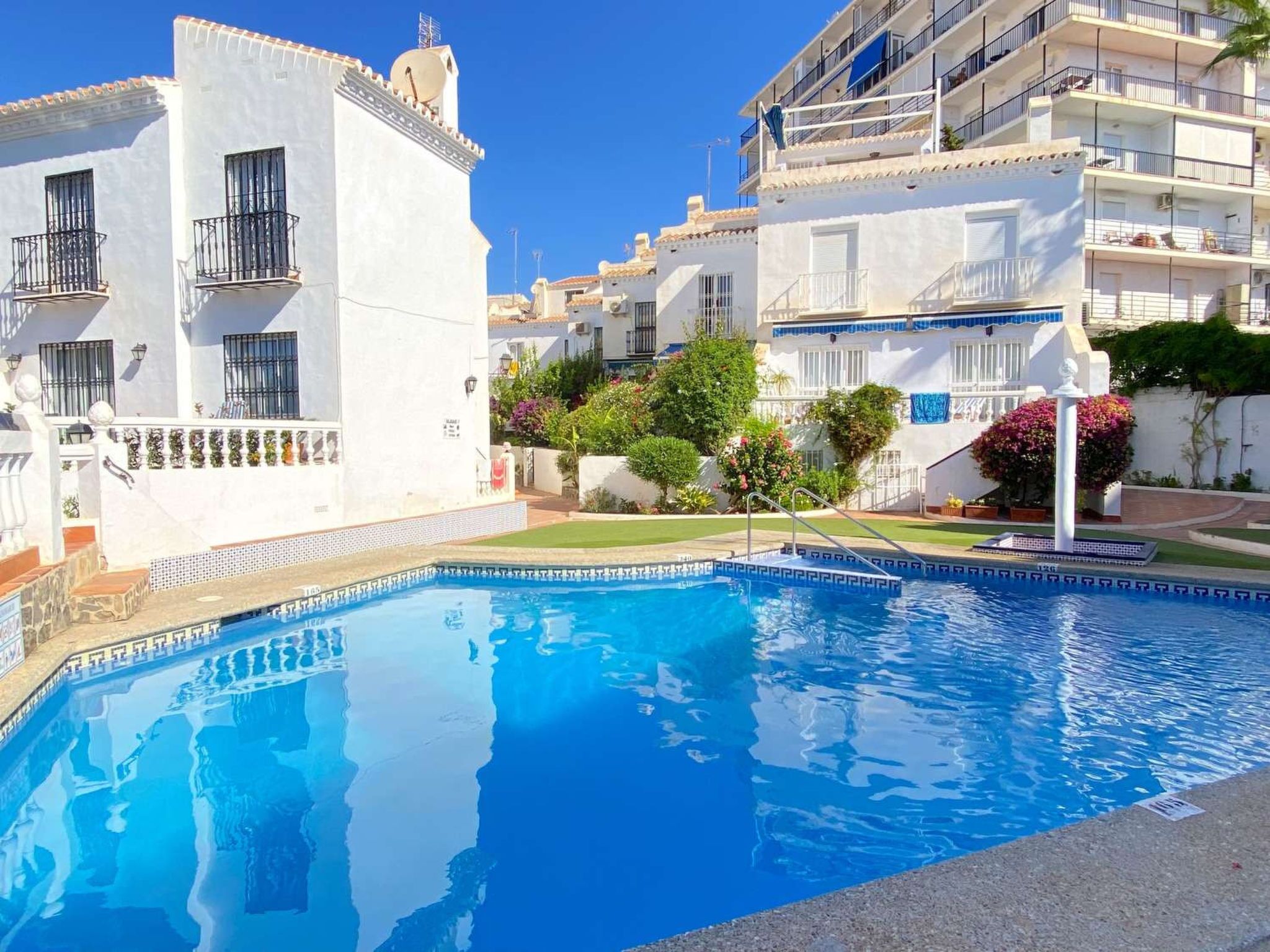 A swimming pool surrounded by white buildings with balconies, a clear blue sky, and some greenery.