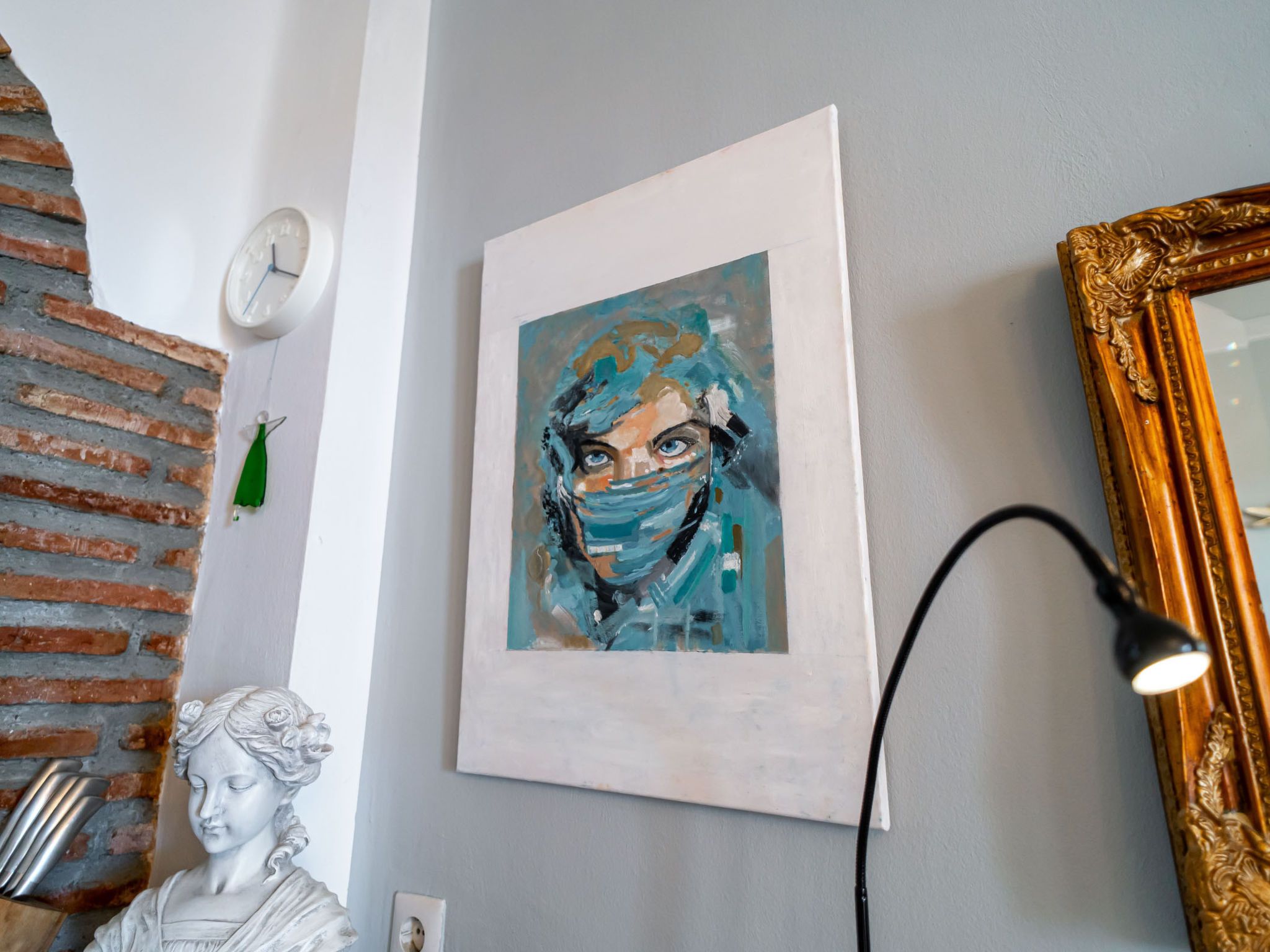 A room with a brick wall, a white clock, a framed artwork, a statue, and a gold-framed mirror.