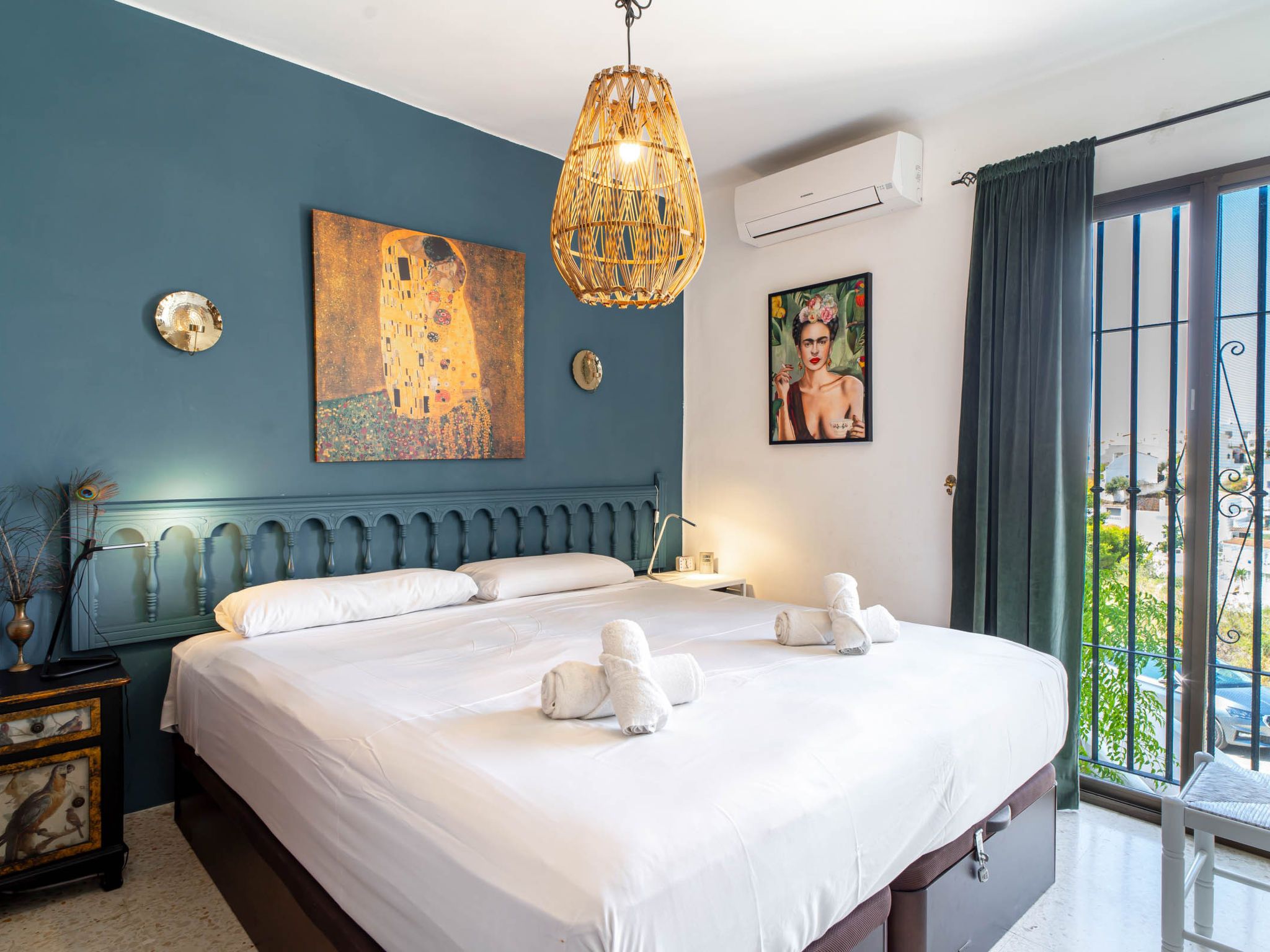 A hotel room with a large bed, a painting on the wall, an air conditioning unit, and a view through the window.