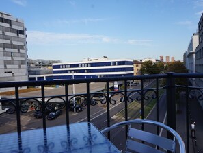 Blick vom Balkon auf einen Parkplatz mit mehreren Autos, ein blau-weißes Gebäude und weitere städtische Strukturen im Hintergrund.