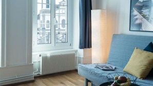 Ein Zimmer mit Blick auf ein Gebäude durch ein Fenster, inklusive Heizkörper, einer Couch mit Kissen und einer Lampe.