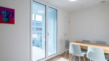 Una habitación con una mesa de madera y cuatro sillas, una puerta corrediza de vidrio que da a un balcón exterior y un cuadro pequeño en la pared.