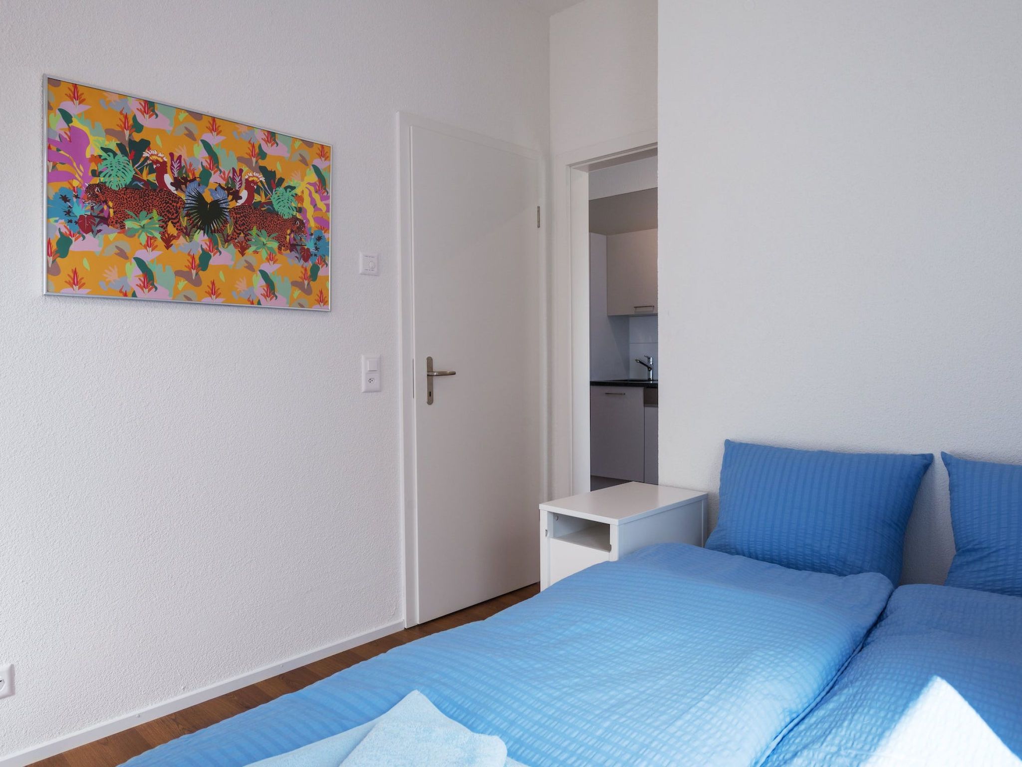 A bedroom with a bed, a nightstand, a door, and a colorful painting on the wall.