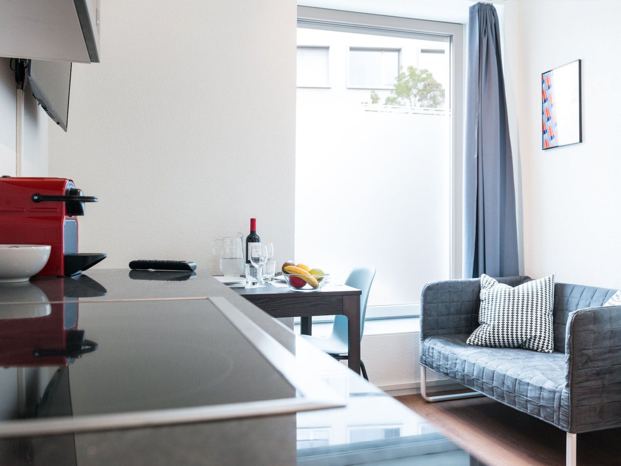 Una cocina moderna con un electrodoméstico rojo, una mesa de comedor con un cuenco de fruta y un sofá gris con un cojín estampado.