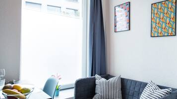 A modern living room with a grey sofa, a glass table with fruits, and a window with curtains.