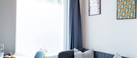 A modern living room with a grey sofa, a glass table with fruits, and a window with curtains.