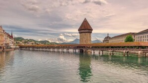 Ein historischer Turm mit einem spitz zulaufenden Dach, eine Stegbrücke und ein ruhiger Fluss, der den Himmel spiegelt.