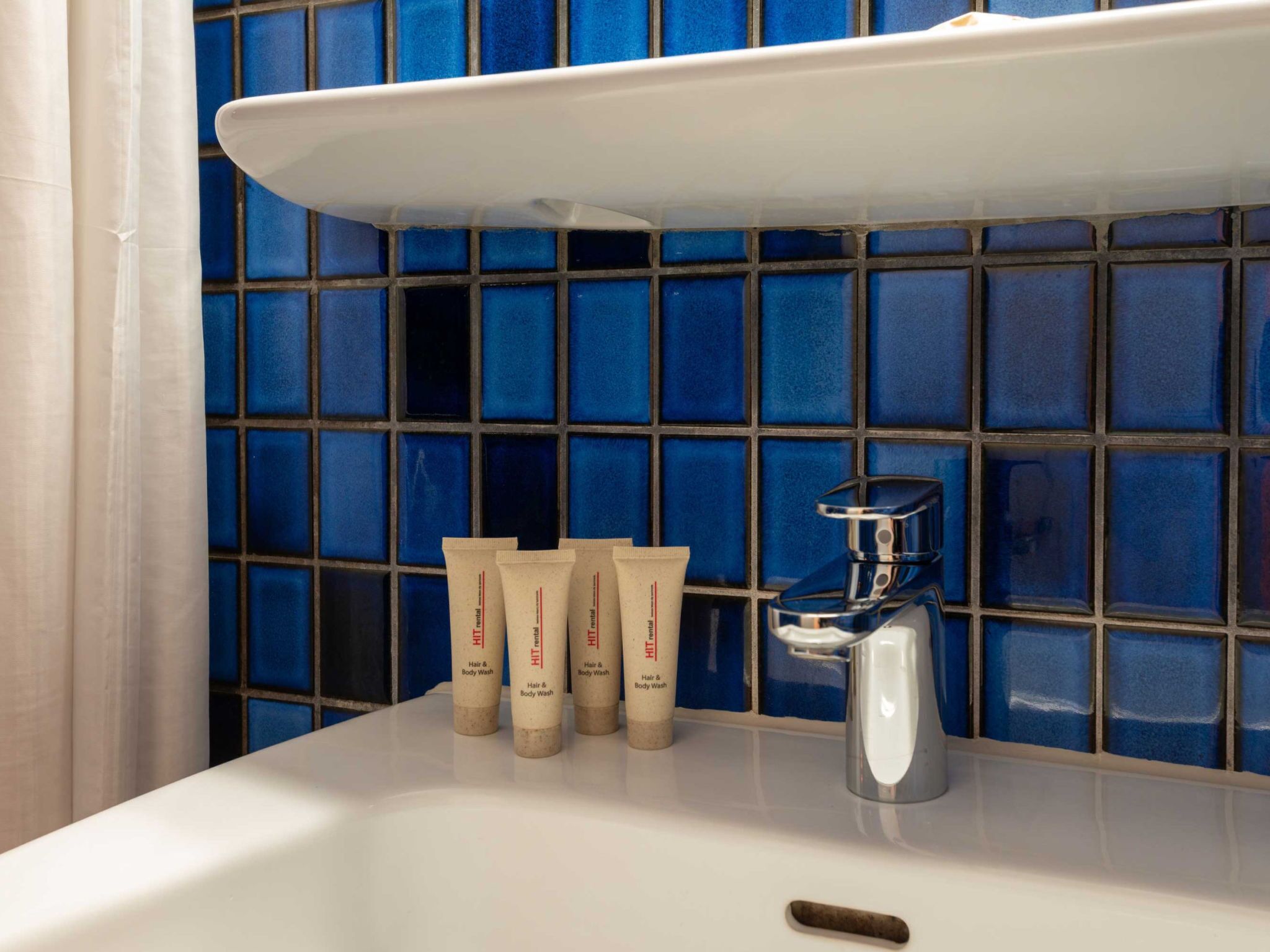 A bathroom with a white sink, a chrome faucet, and a blue tiled wall. There are three tubes of hair and body wash on the sink.