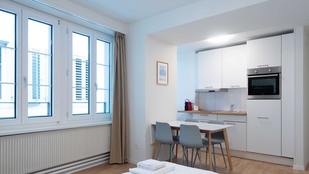 Ein modernes, kompaktes Hotelzimmer mit großem Fenster, einer Essecke und einer Küchenzeile.