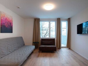 Une pièce avec un canapé gris, un fauteuil marron, une petite table, un téléviseur fixé au mur et une fenêtre avec des rideaux.