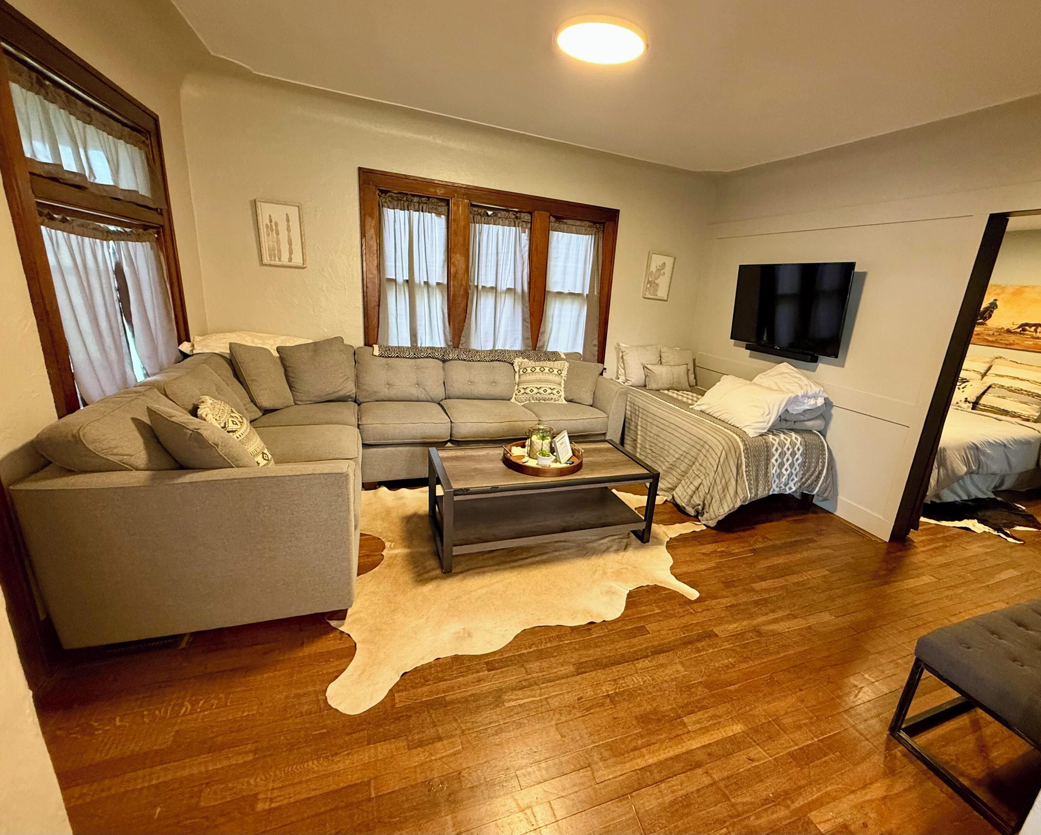 A living room with a sectional sofa, a small coffee table, a bed with a patterned bedspread, and a mounted TV.
