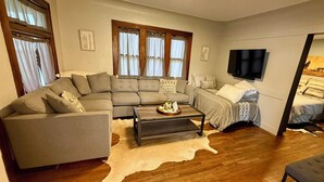 A living room with a sectional sofa, a small coffee table, a bed with a patterned bedspread, and a mounted TV. - 1-4 Guests, 1 Bath Farm Stay at a Pygmy Goat Farm in Byron Center, Michigan (Byron Center)