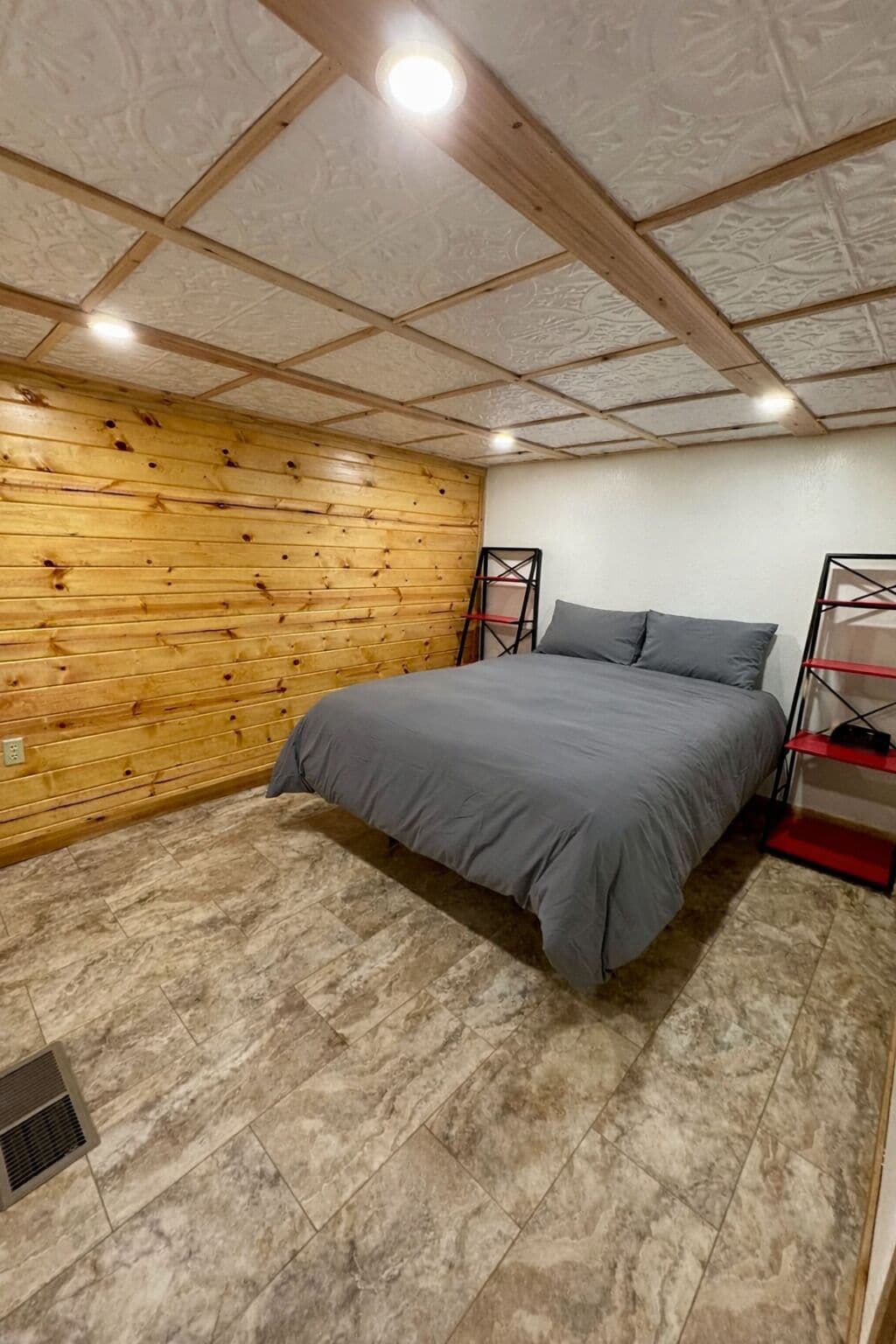 Ein Schlafzimmer mit einer Holzwand, einem Bett mit grauer Bettwäsche und einem Regal in Rot und Schwarz.