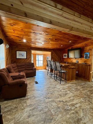 A log cabin interior with a living area, a kitchen with bar seating, and a television.