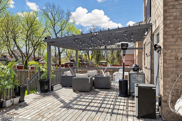 Un patio techado con muebles de exterior, una pérgola con luces colgantes y un edificio de ladrillo al fondo.