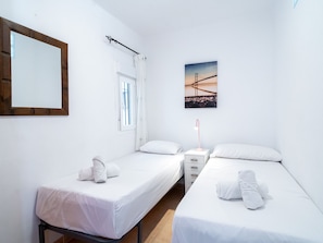 Un dormitorio con dos camas individuales, una mesita de noche, una lámpara, una ventana con cortinas, un espejo montado en la pared y un cuadro de un puente.