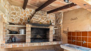 A stone and brick outdoor kitchen with a built-in grill, shelves with cooking utensils, and a stone floor. - Finca Can Nin by Interhome (Santanyí)