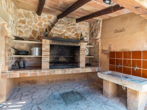 A stone and brick outdoor kitchen with a built-in grill, shelves with cooking utensils, and a stone floor. - Finca Can Nin by Interhome (Santanyí)