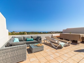Una terraza en la azotea con sofá, sillones de lounge y una mesa, con vistas a un paisaje costero.