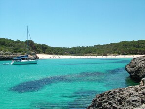 Eine Küstenlandschaft mit einem Segelboot in der Nähe einer felsigen Küste, einem Sandstrand und einer bewaldeten Hügelkette im Hintergrund.