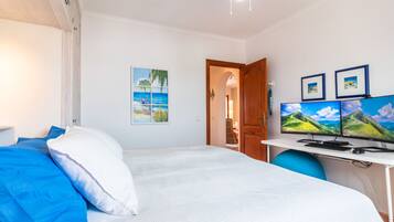 A bedroom with a bed, a desk with two monitors, a ceiling fan, and a door leading to another room.