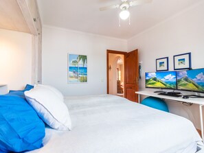 Un dormitorio con cama, un escritorio con dos monitores, un ventilador de techo y una puerta que da a otra habitación.