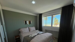 A bedroom with a bed, a nightstand, a window with curtains, and a wall-mounted coat rack. - Clócio 201 - Beachfront apartment on Cabo Branco beach (João Pessoa)