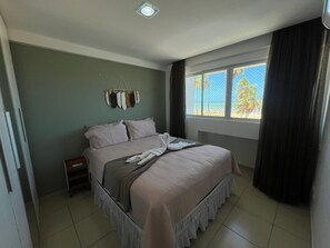 A bedroom with a bed, a nightstand, a window with curtains, and a wall-mounted coat rack. - Clócio 201 - Beachfront apartment on Cabo Branco beach (João Pessoa)