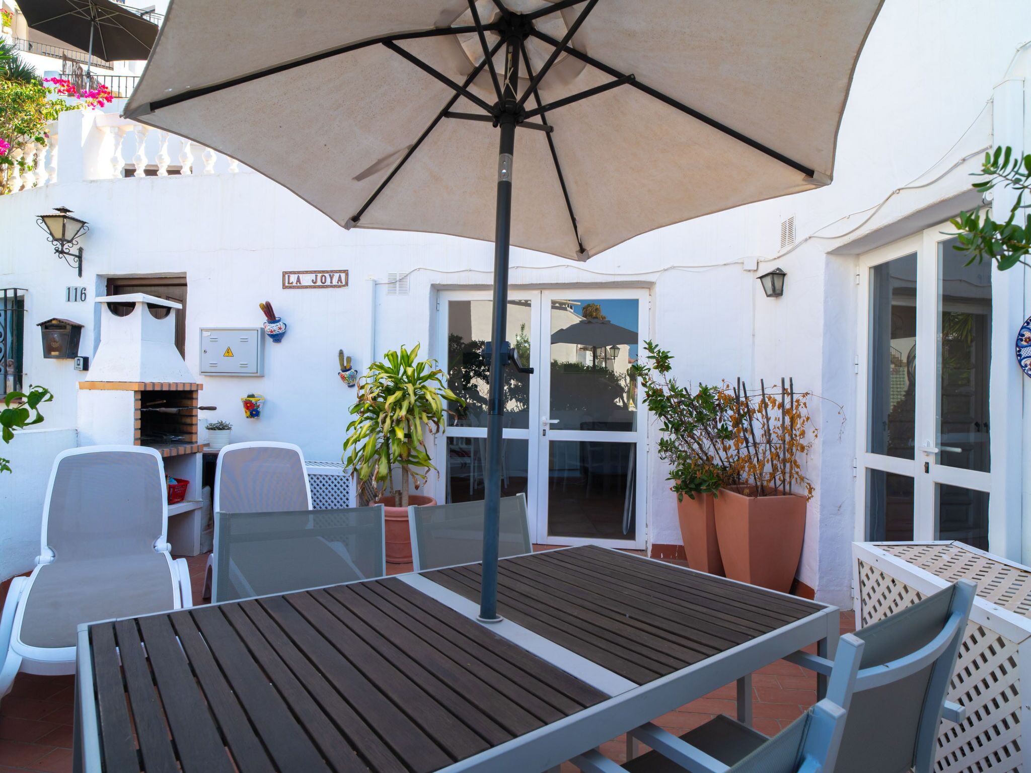 An outdoor patio with a table and chairs, a large umbrella, potted plants, and a sign reading 'LA JOYA'.