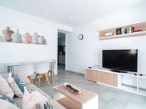A modern living room with a flat-screen TV, a dining area with chairs, a sofa with cushions, and a wall-mounted shelf with books. - Apartment  by Interhome (Nerja)