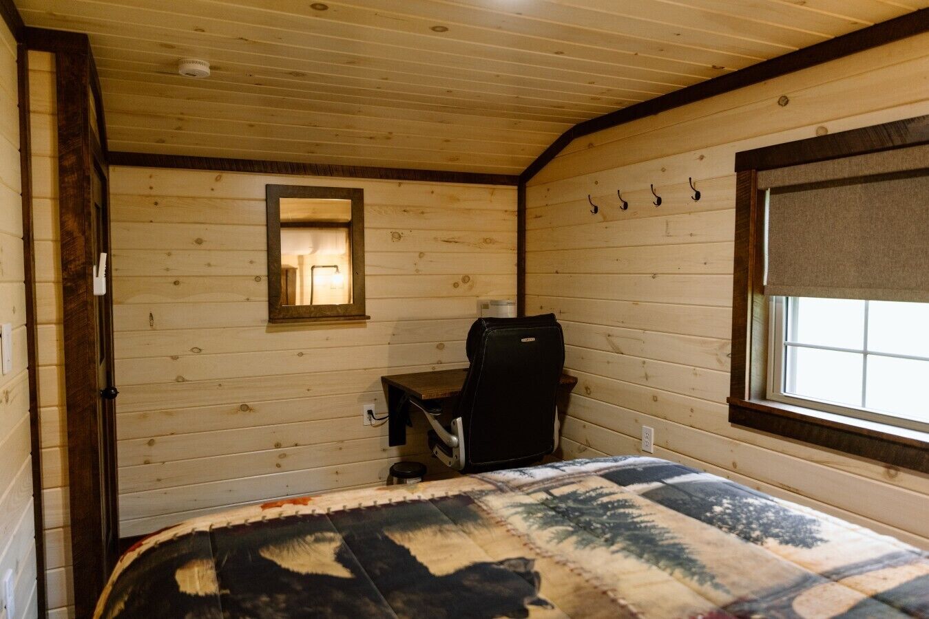 A room with wooden walls, a bed with a patterned blanket, a desk with a chair, a window with a blind, and a mirror on the wall.