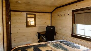 A room with wooden walls, a bed with a patterned blanket, a desk with a chair, a window with a blind, and a mirror on the wall.