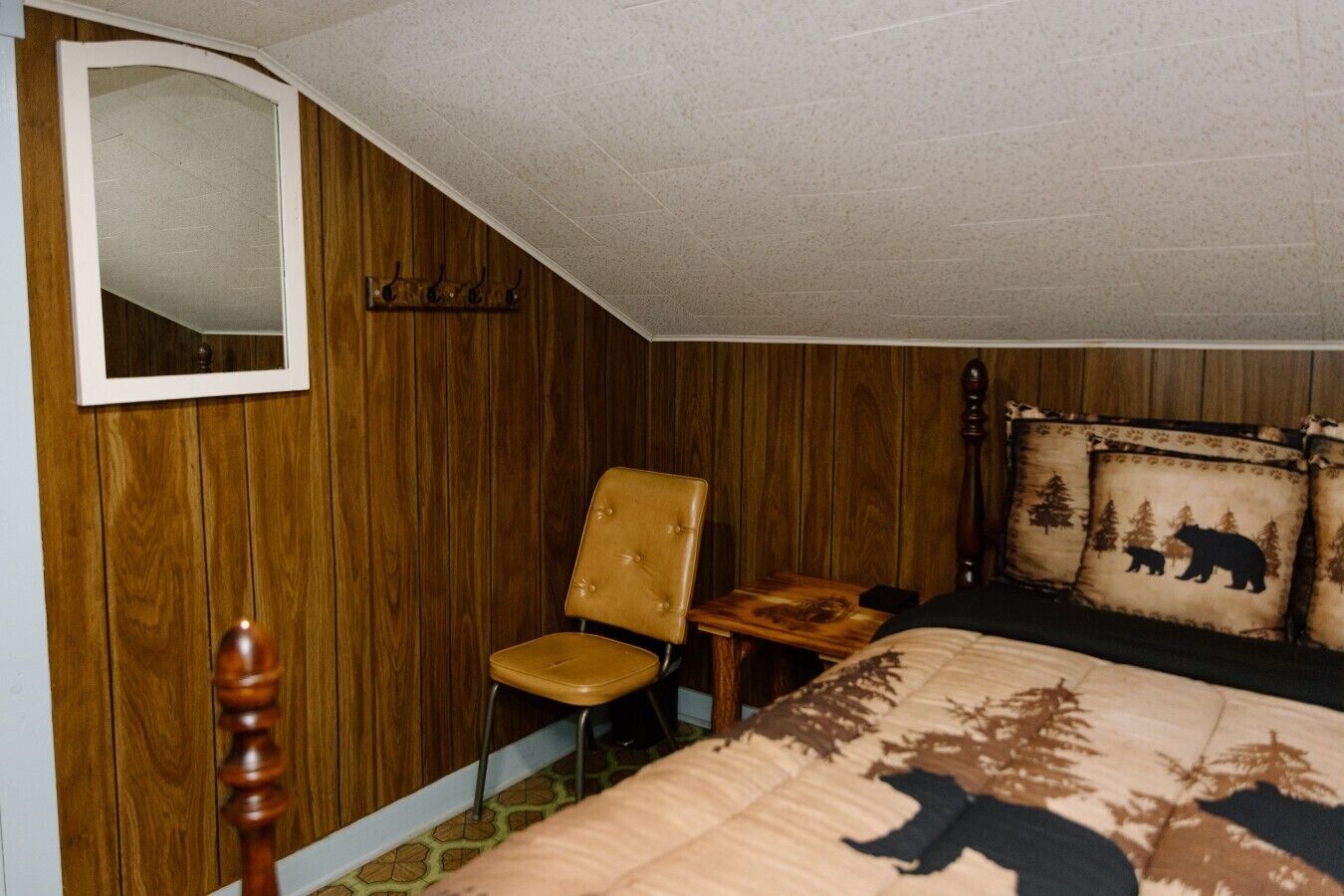 Un dormitorio con pared revestida de madera, un espejo, un sillón capitonado, una mesita de noche, una cama con un cojín con motivos de oso y una lámpara de pie de madera.