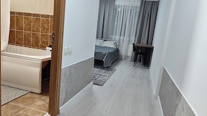 Un pasillo estrecho que conduce a un dormitorio con una cama, una mesita y una silla. Hay un baño con bañera y zona de ducha.
