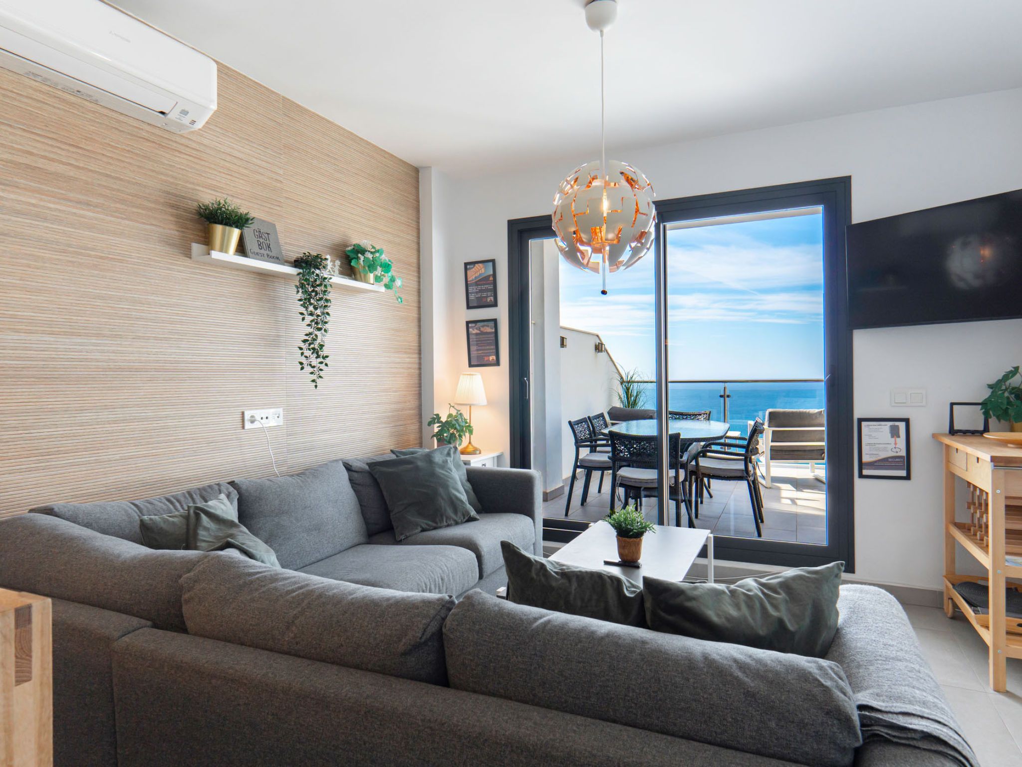 Un salon moderne avec un canapé gris, une table à manger avec des chaises, un luminaire suspendu et une vue sur la mer à travers de grandes portes vitrées.