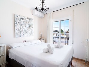 Une chambre d’hôtel avec un grand lit, un ventilateur de plafond, une fenêtre donnant sur des immeubles et un tableau au mur.