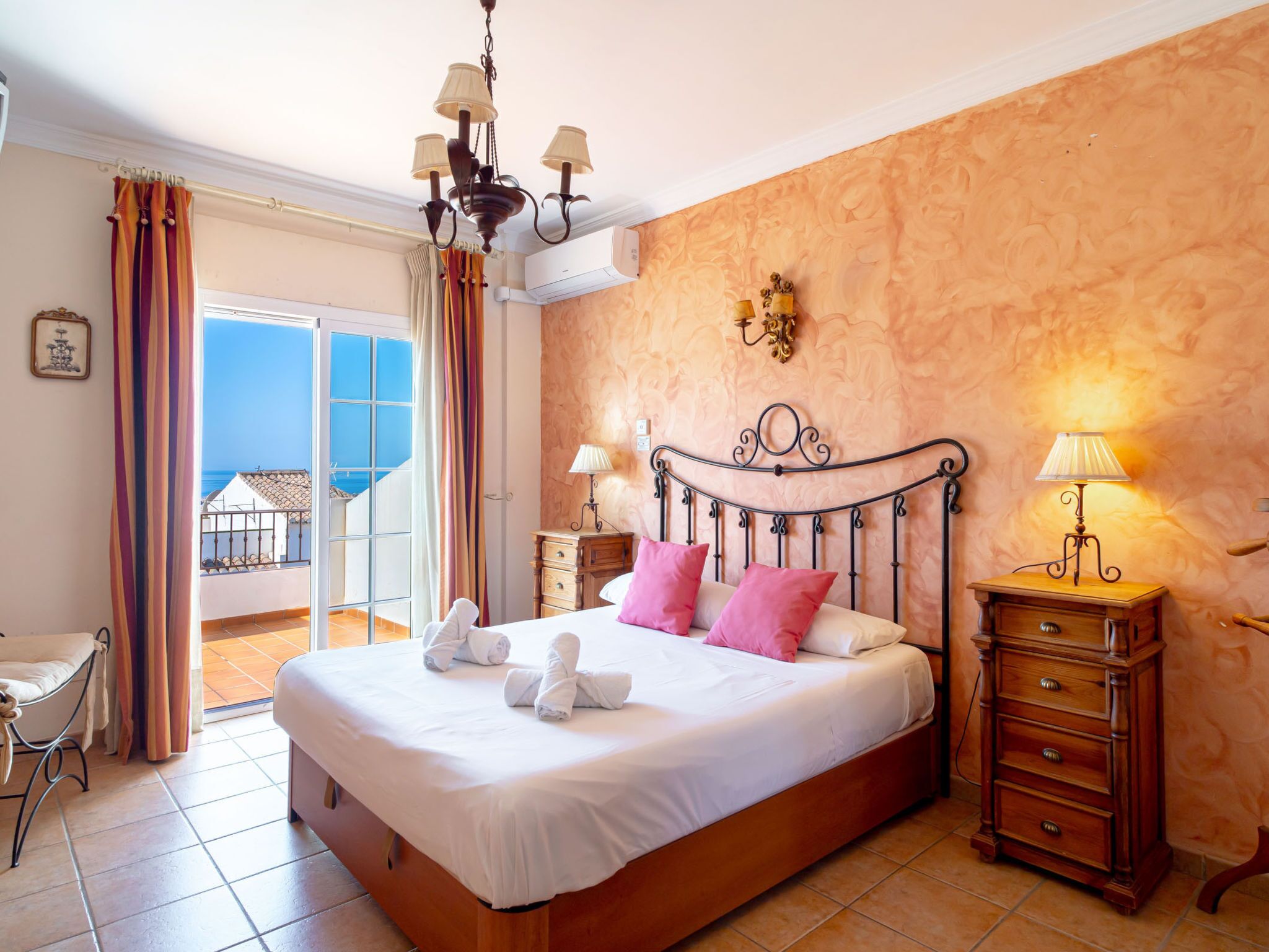 A hotel room with a bed, a wooden nightstand, a wall-mounted lamp, and a view of the sea through a balcony door.